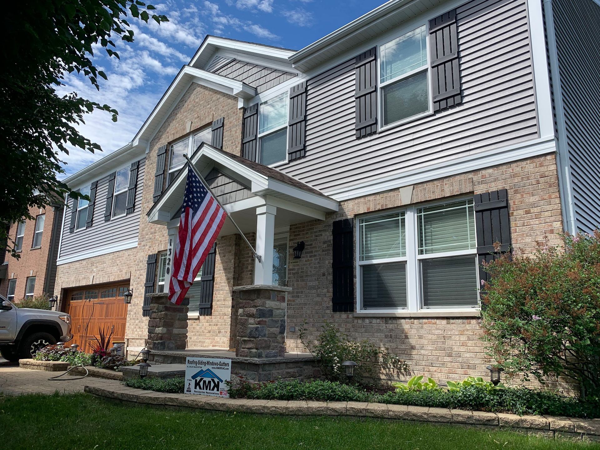 A two-story suburban house with a brick and gray siding exterior, featuring a stone-column porch and an American flag.