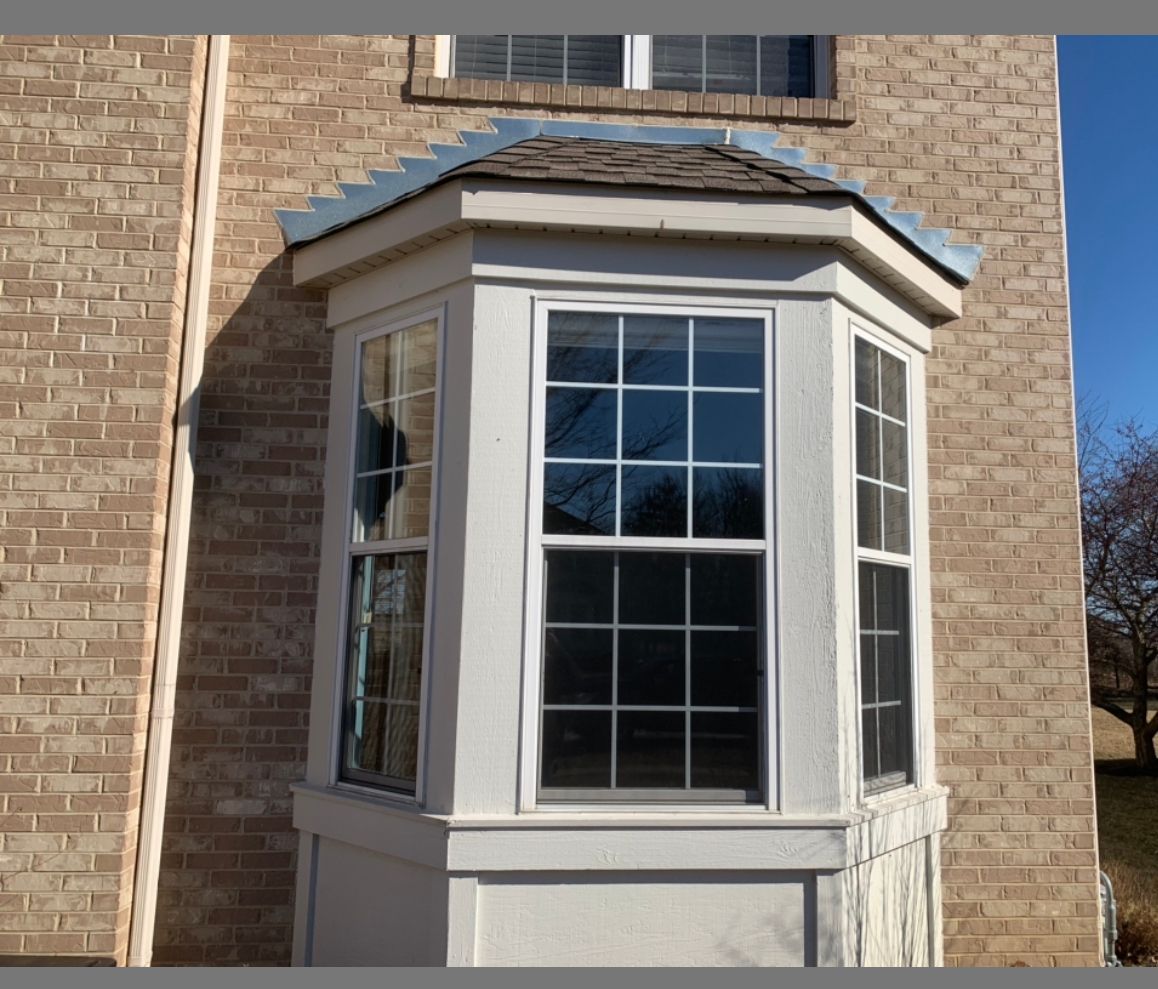 A white bay window with a grid pattern, installed in a tan brick wall under a small shingled roof.