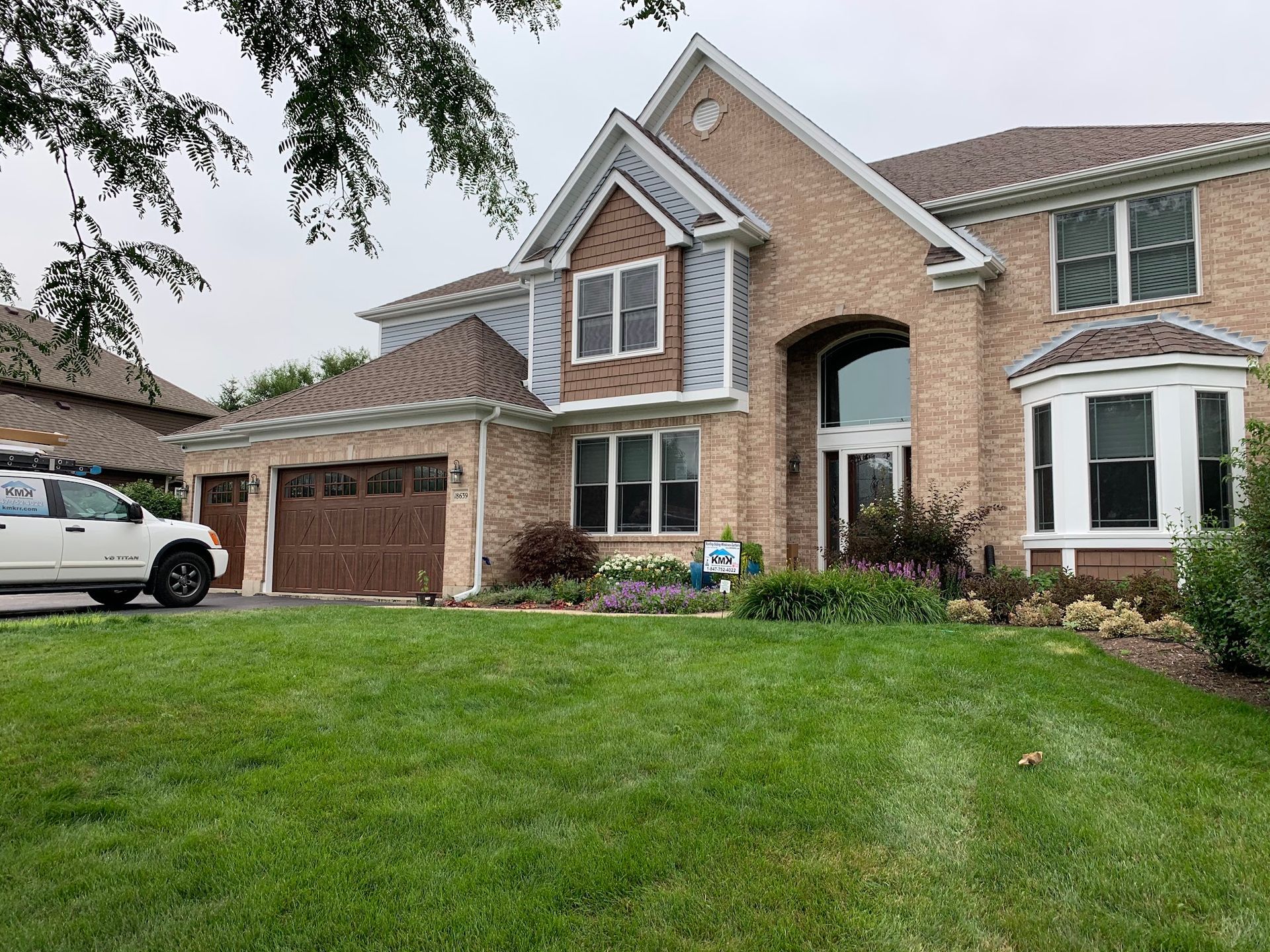 A large, two-story tan brick suburban house with a brown garage and a green lawn under a cloudy sky.