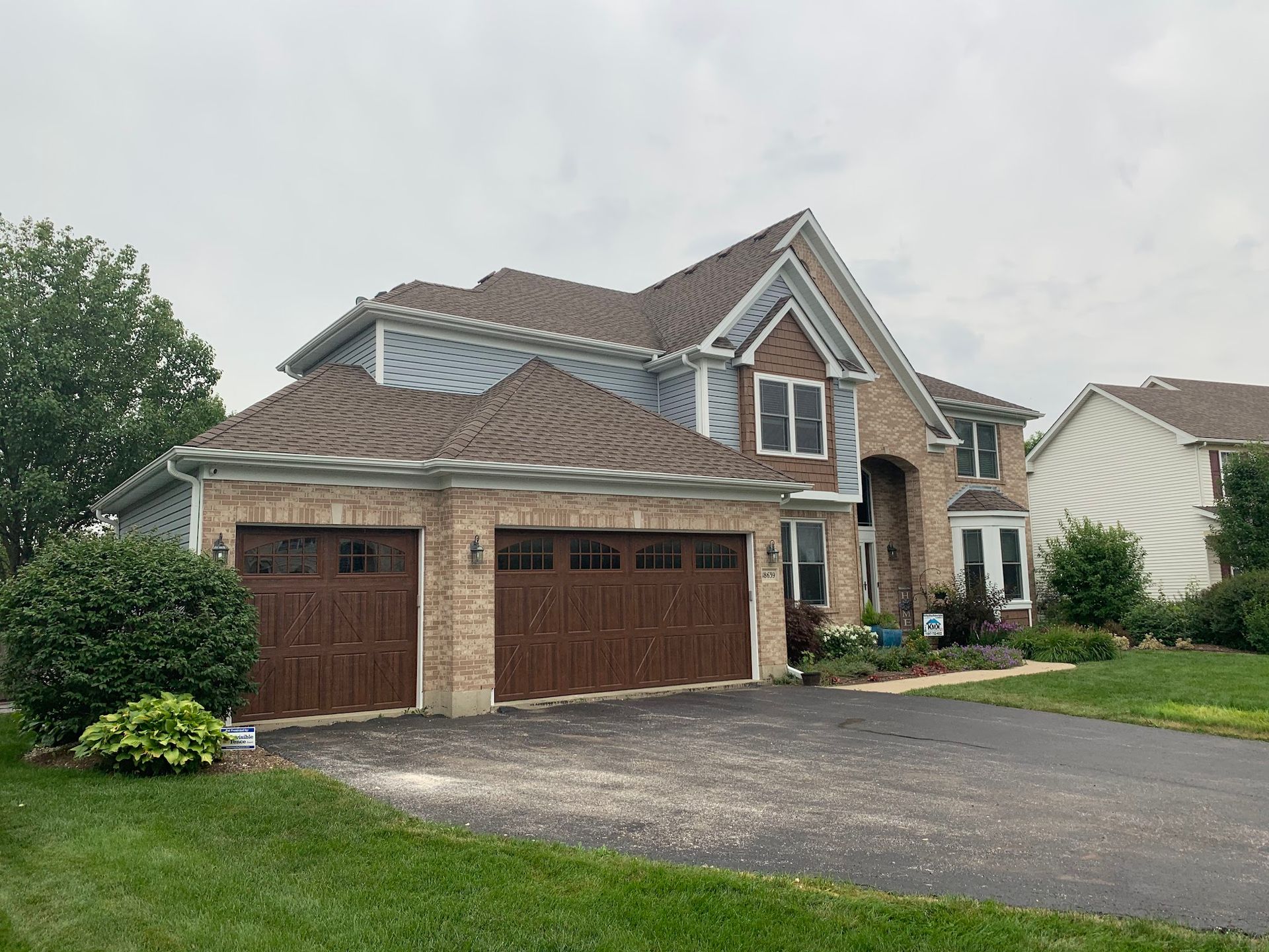 A two-story brick suburban house with a three-car garage and asphalt driveway on an overcast day.