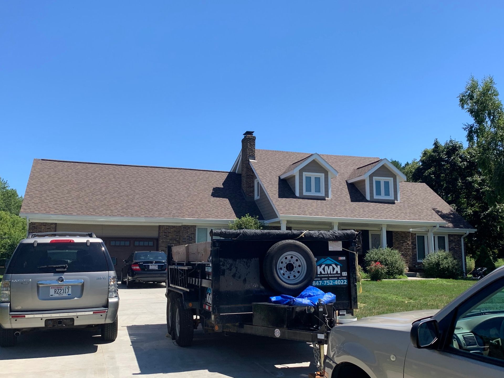 A house with a newly shingled roof, a chimney, and two dormers, with vehicles and a dump trailer in the driveway.