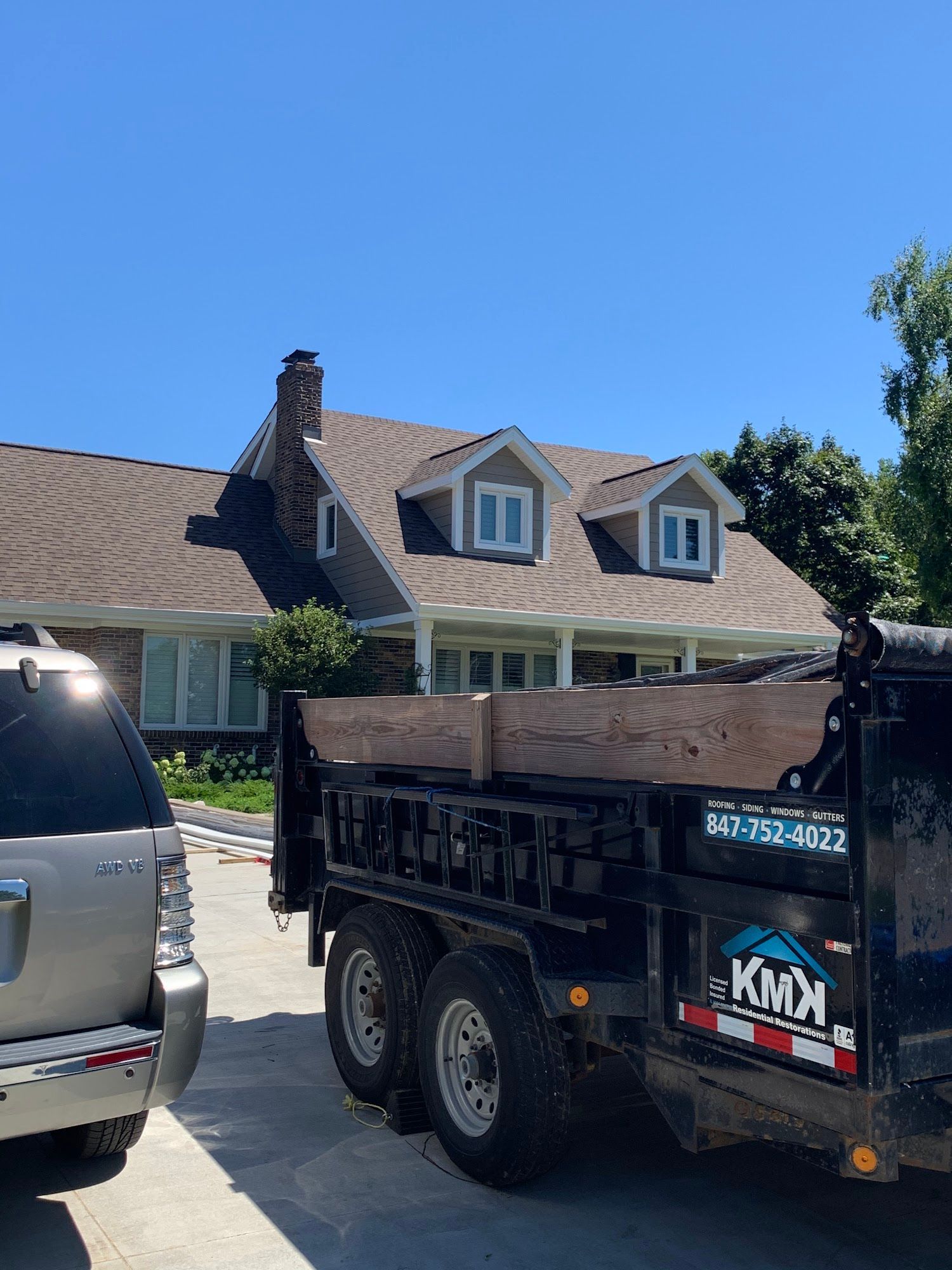 A black dump trailer parked in a driveway in front of a house with brown shingles and two dormer windows under a blue sky.
