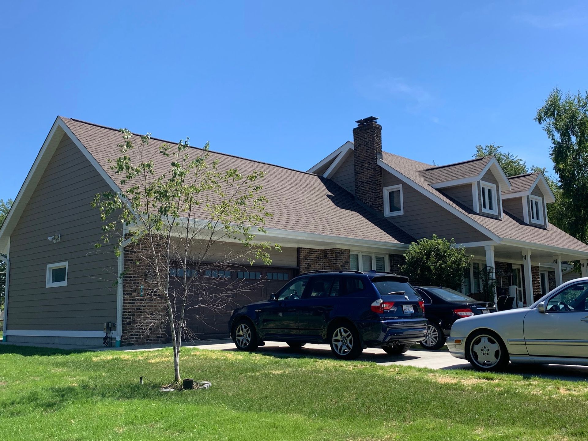 A tan house with a brick garage facade, two cars parked in the driveway, a chimney, and a small tree on a sunny day.