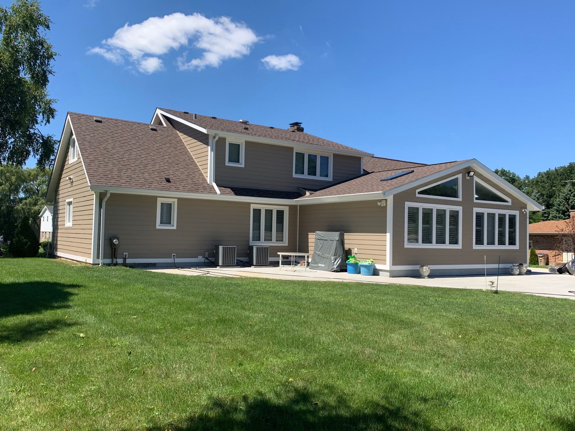 A two-story, tan-sided house with a dark brown roof and a sunroom extension, set against a green lawn and blue sky.