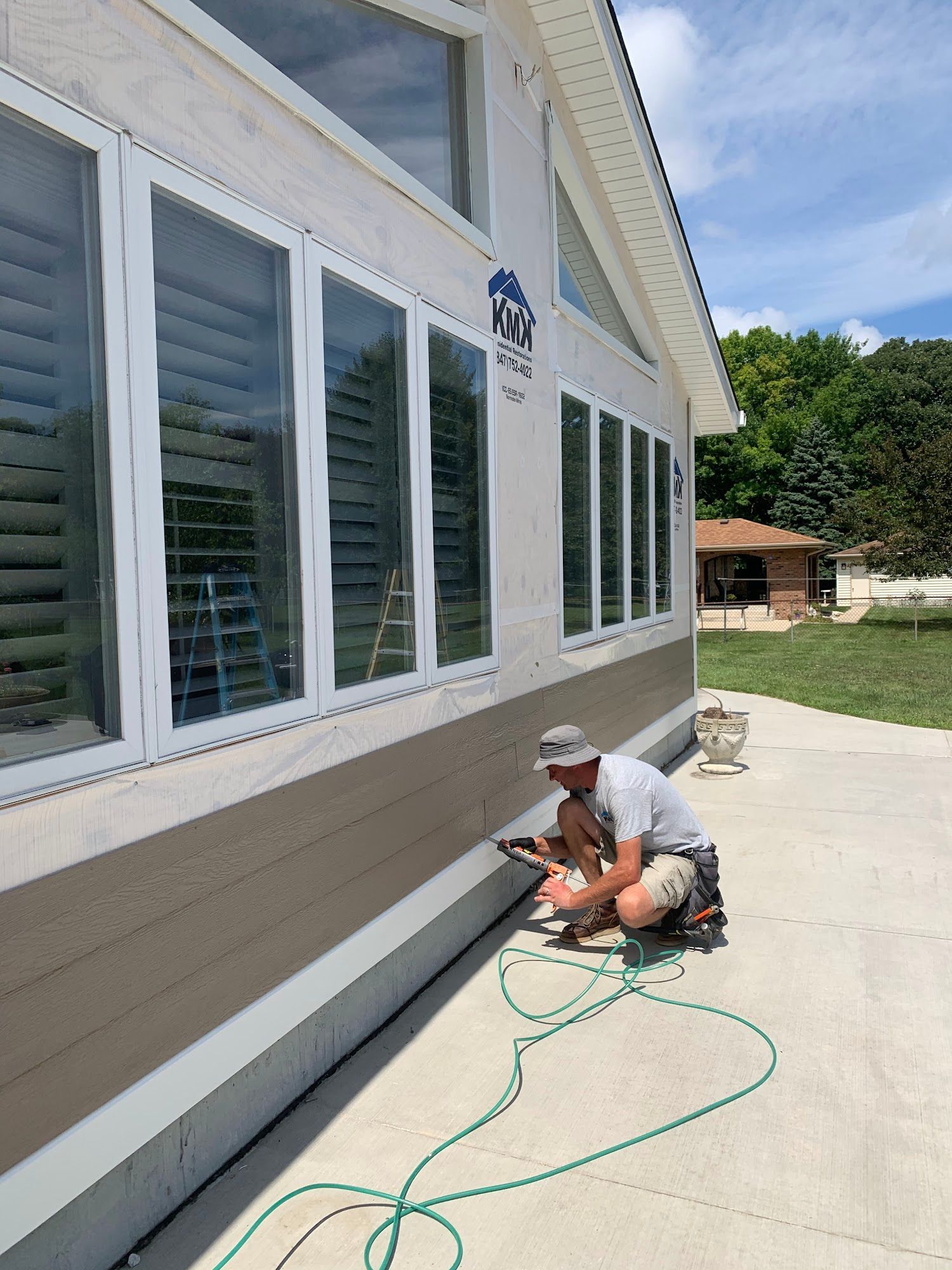 A worker kneels on a concrete patio, using a nail gun to install tan horizontal siding on a house wall beneath windows.