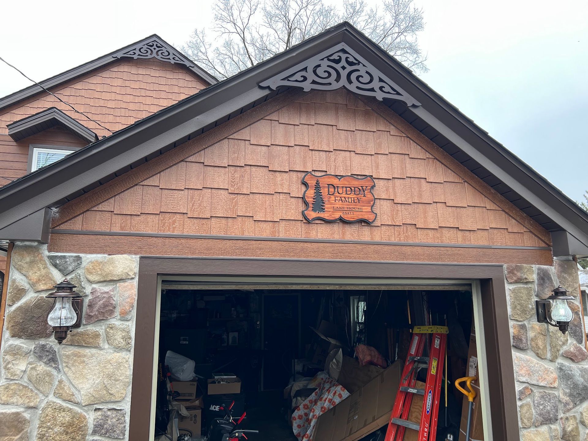 The front of a home with a garage featuring stone walls, brown cedar shake shingles, and a wooden decorative sign.