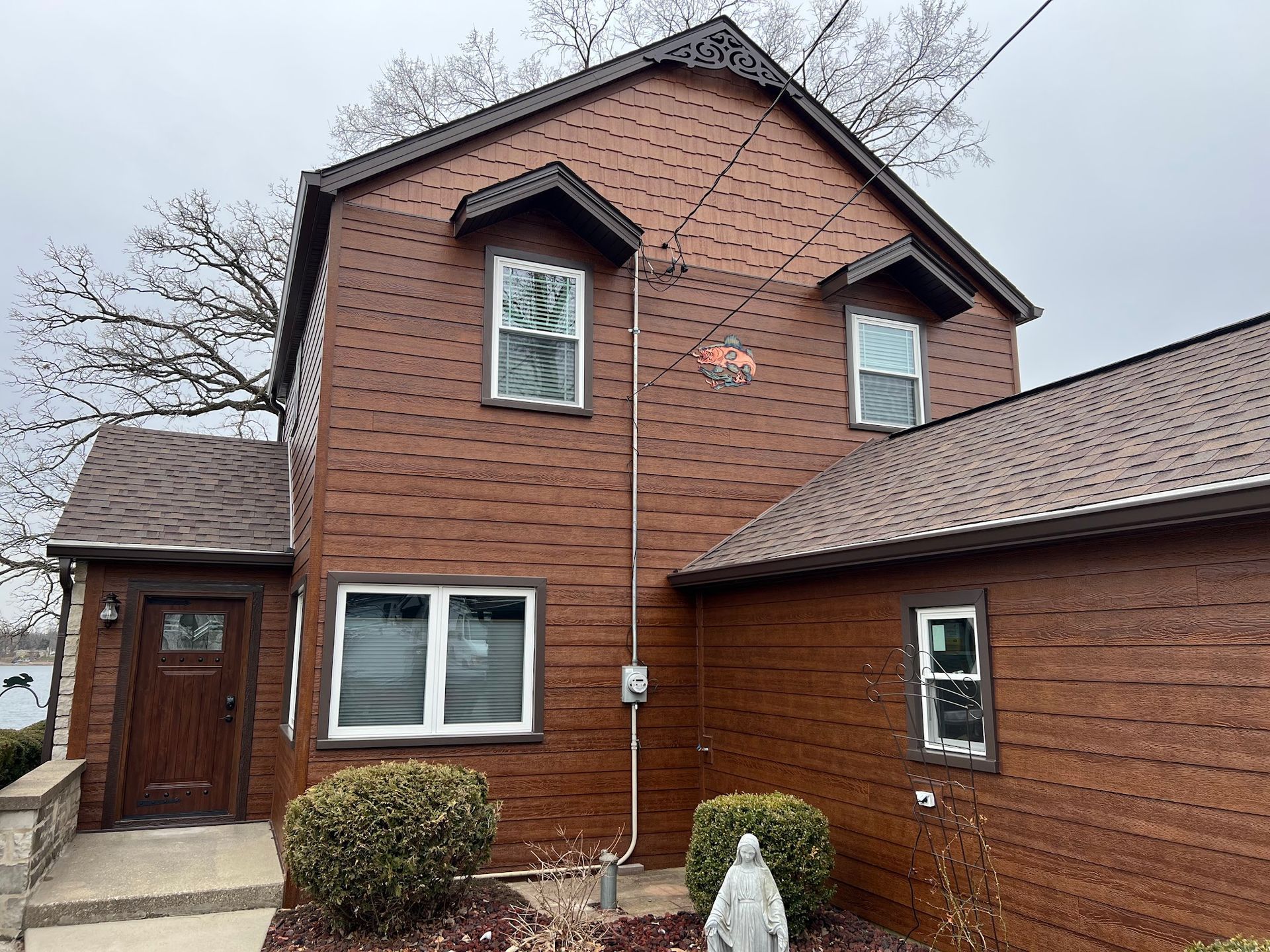 A two-story house with brown textured siding, multiple windows, a brown front door, and a gray shingled roof under a sky.