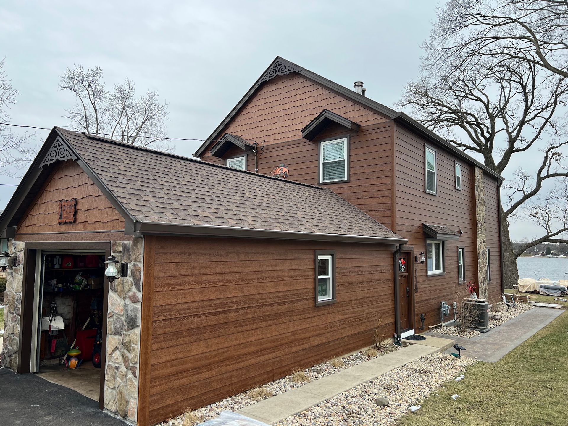 A two-story house with dark brown wood-look siding, stone accents, and an attached garage under a gray sky.