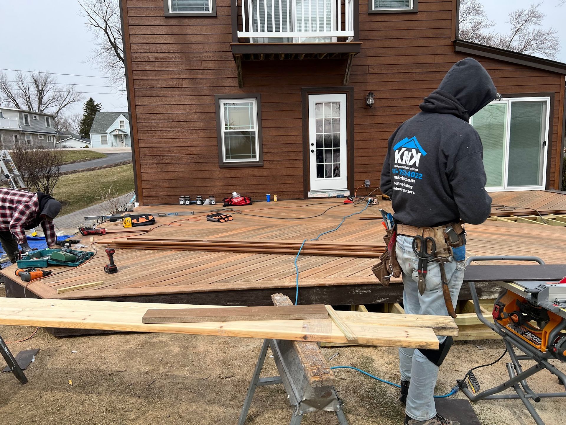 Two people work on a wooden deck project in front of a two-story brown house under a cloudy sky.
