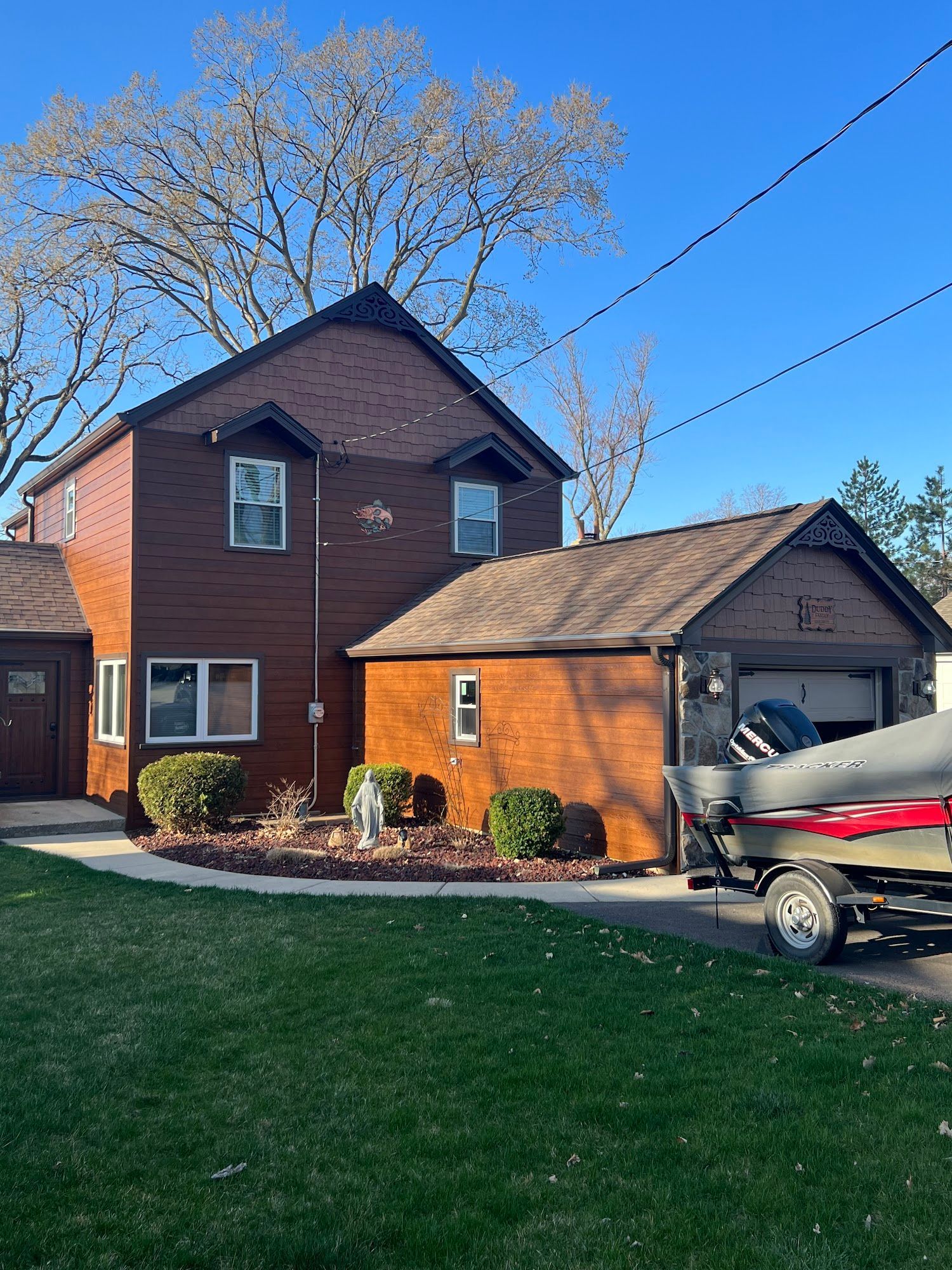 A two-story brown house with a garage and a boat parked on a trailer in the driveway under a clear blue sky.