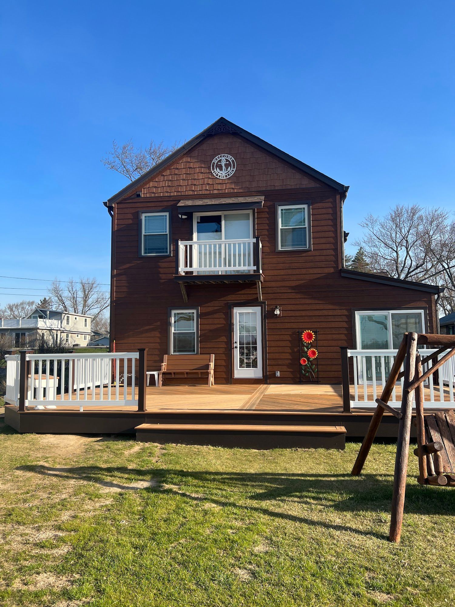 A two-story brown house with a large wooden deck, white railings, a second-floor balcony, and a grassy yard.