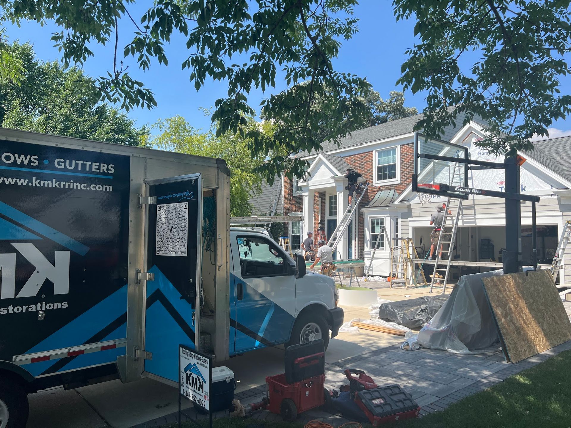 A work truck parked in a driveway in front of a brick house during exterior renovations, with ladders and tools visible.