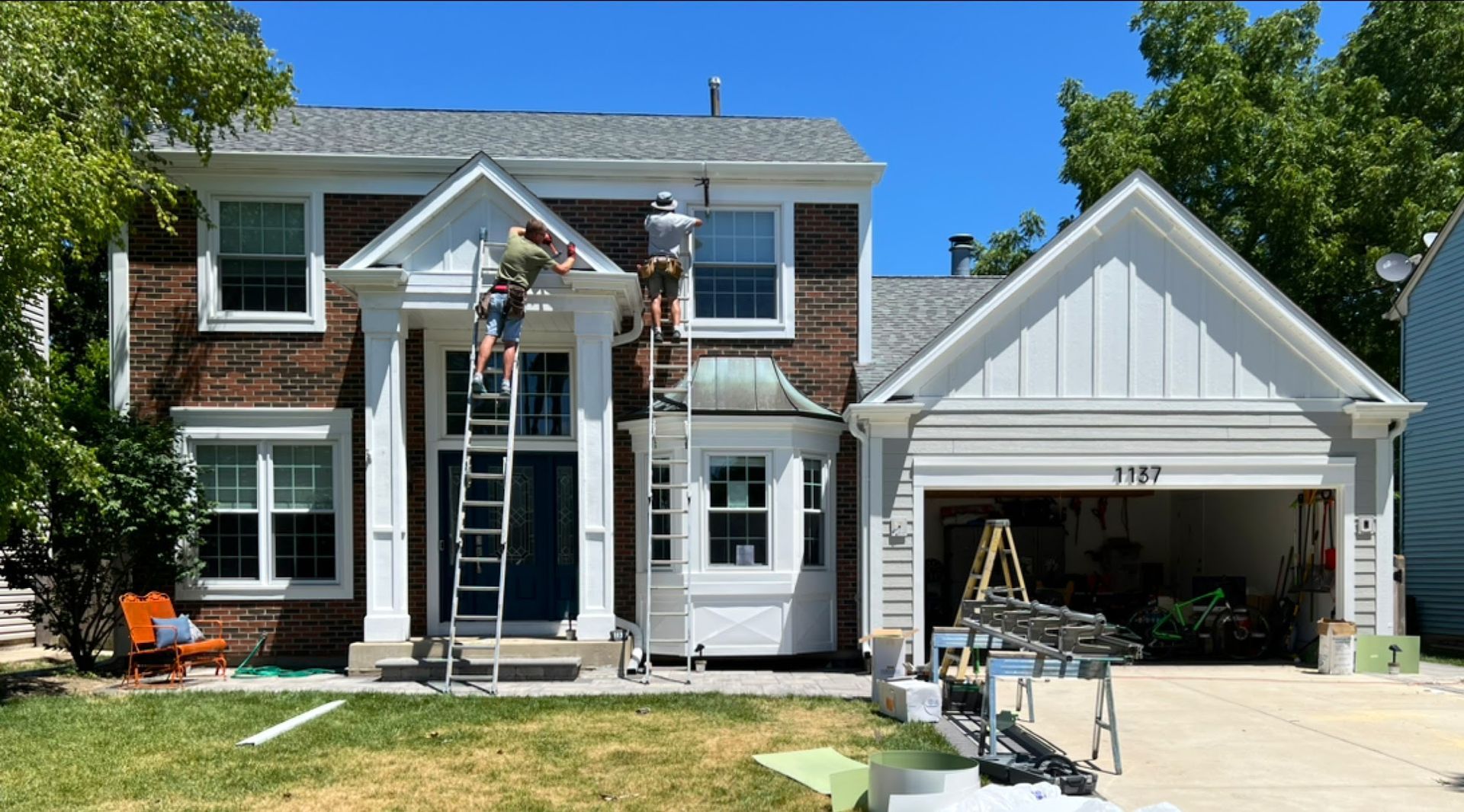 Two people on ladders perform maintenance on the exterior trim of a brick two-story house with an attached garage.