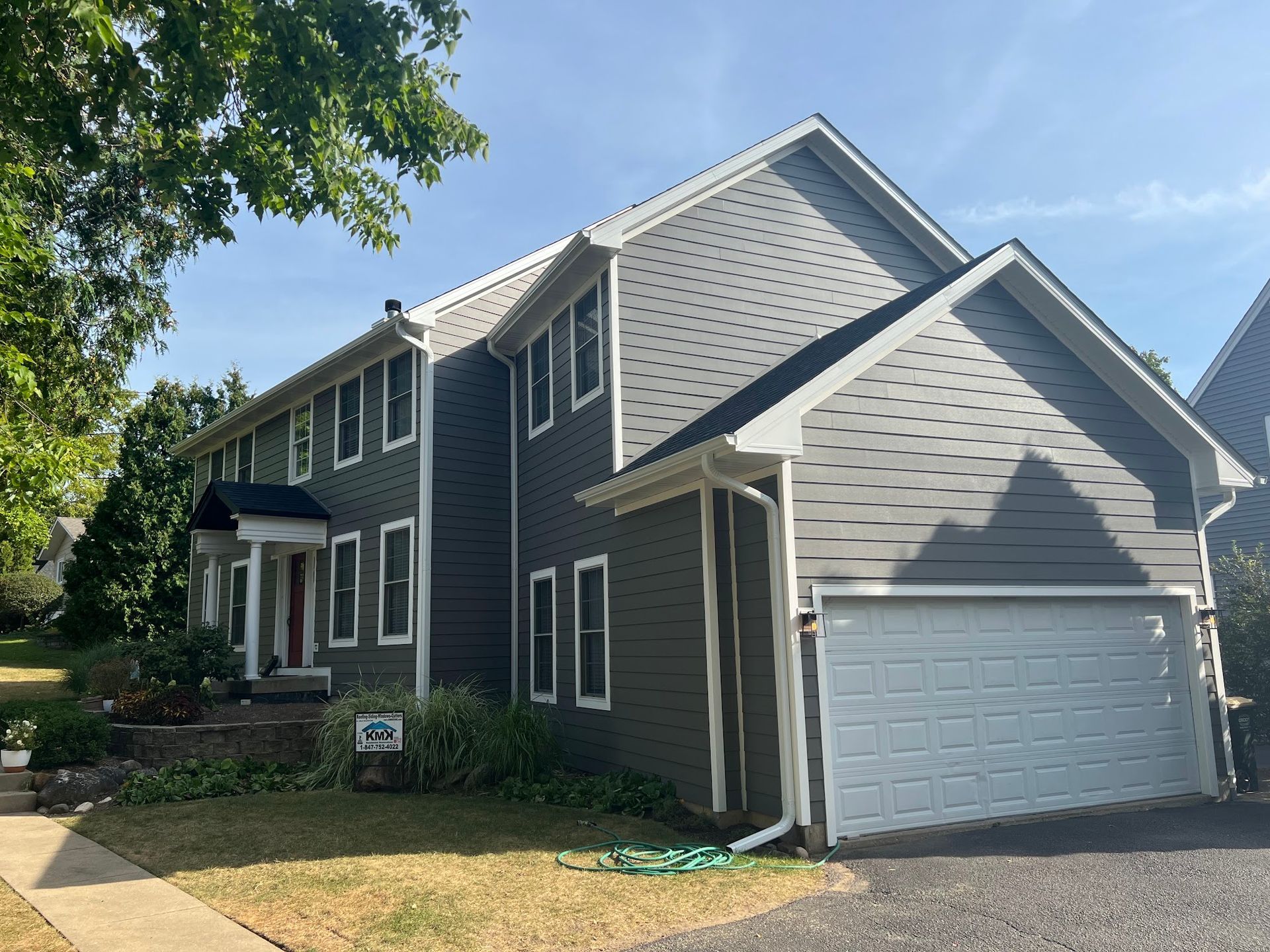 Two-story house with dark gray siding, white trim, a front porch, and an attached two-car garage under a sunny blue sky.