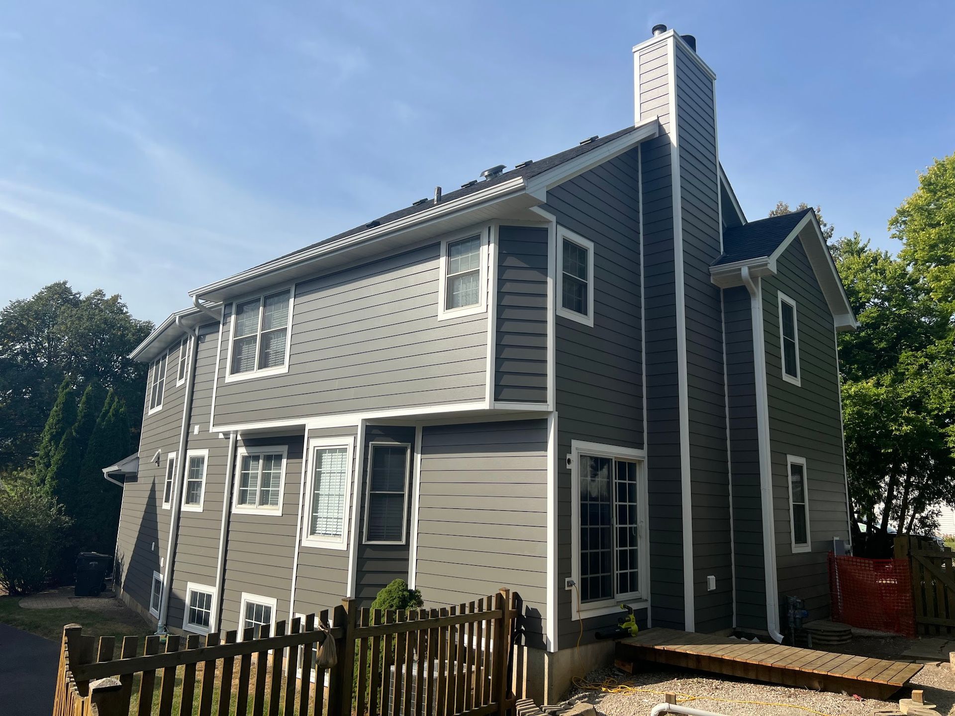 A gray two-story house with white trim and a prominent chimney on the side, seen from a backyard corner.