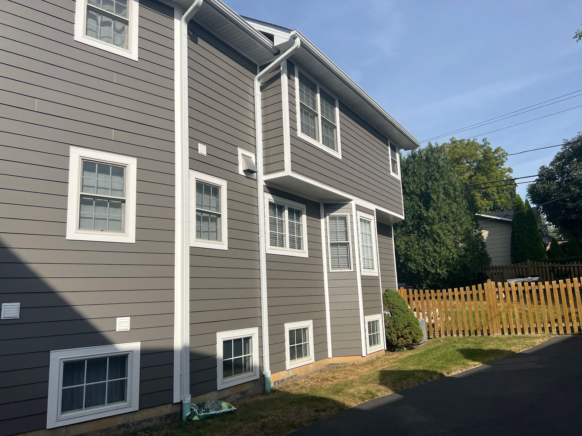 A gray, two-story house with white trim and multiple windows, viewed from the side with a wooden fence in the background.