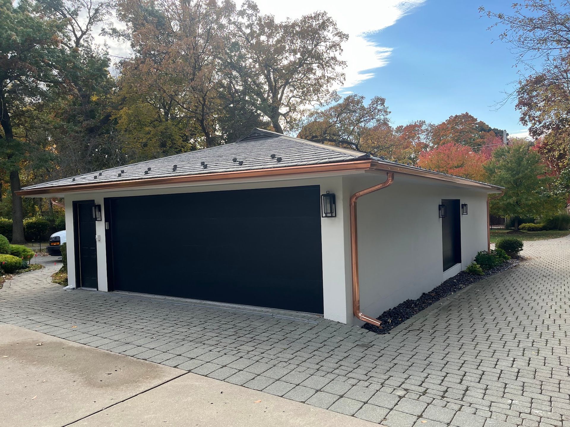 Modern white garage with a black door, dark-tiled roof, and copper gutters on a paved driveway surrounded by autumn trees.