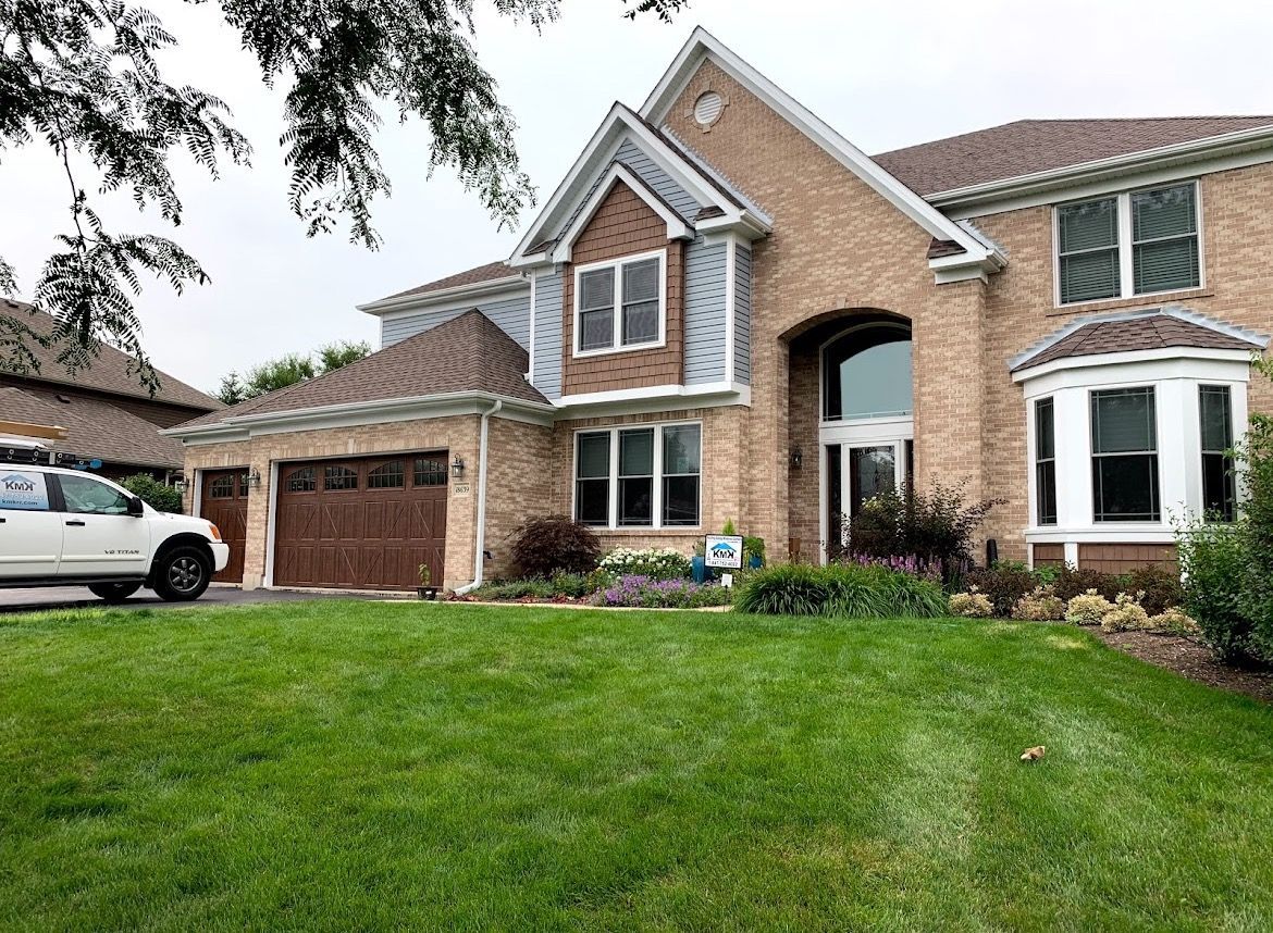 A large, two-story tan brick suburban house with a multi-car garage and a white car parked in the driveway.
