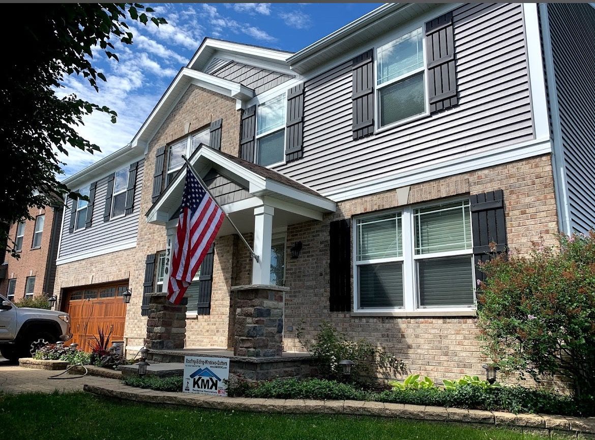 A two-story house with tan brick, grey siding, black shutters, and an American flag hanging by the front porch.