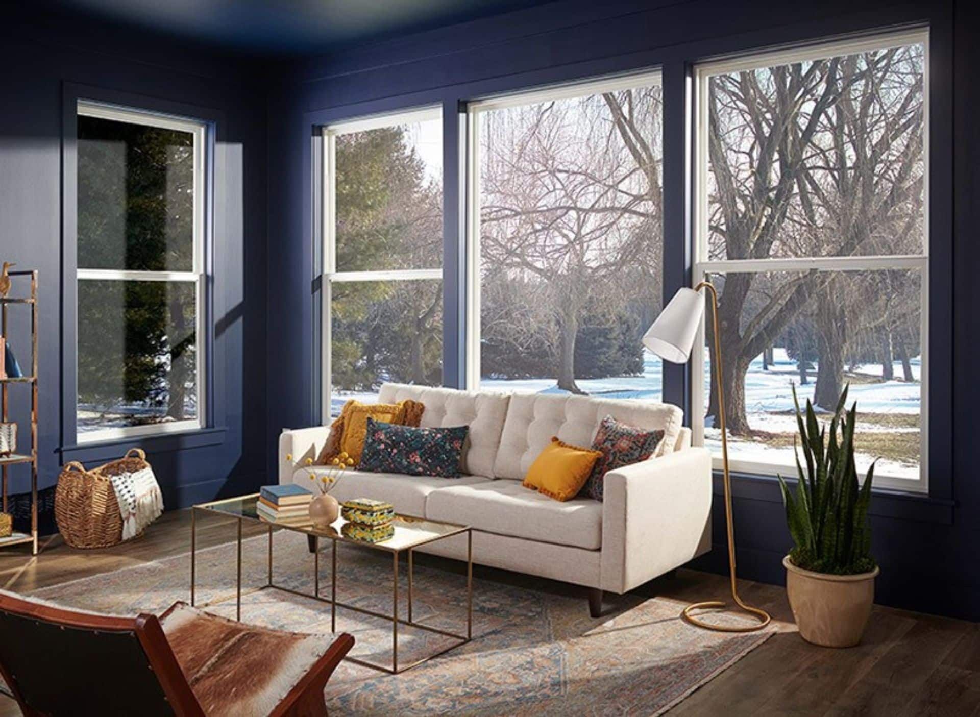 A modern living room with deep blue walls, a white sofa, a gold coffee table, and large windows overlooking trees.