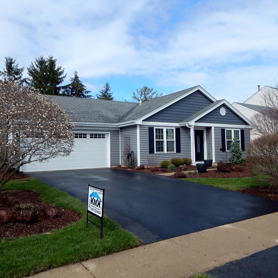 A gray, single-story ranch house with a white garage door, black shutters, and a front yard sign on a paved driveway.