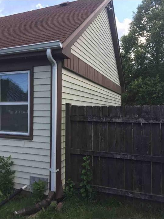 A house exterior with cream siding, brown trim, a white gutter downspout, and a wood fence in the yard.