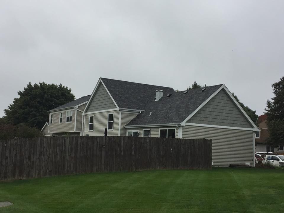 A two-story, light-colored suburban house with dark gray shingles and a wooden privacy fence, viewed from a grassy yard.