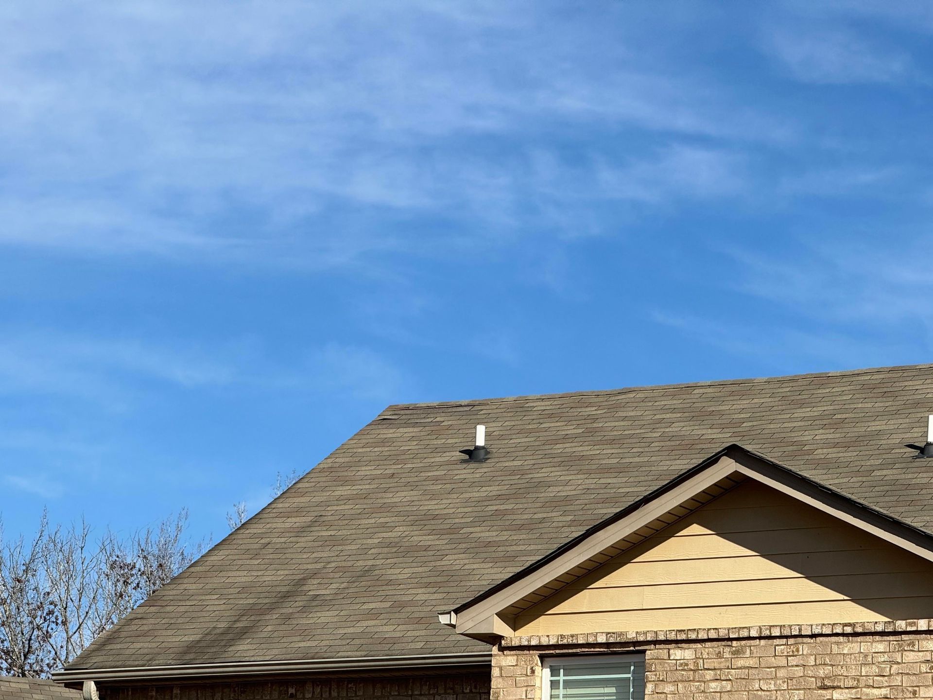 A house roof with shingles and two white plumbing vent pipes against a blue sky with light, wispy clouds.