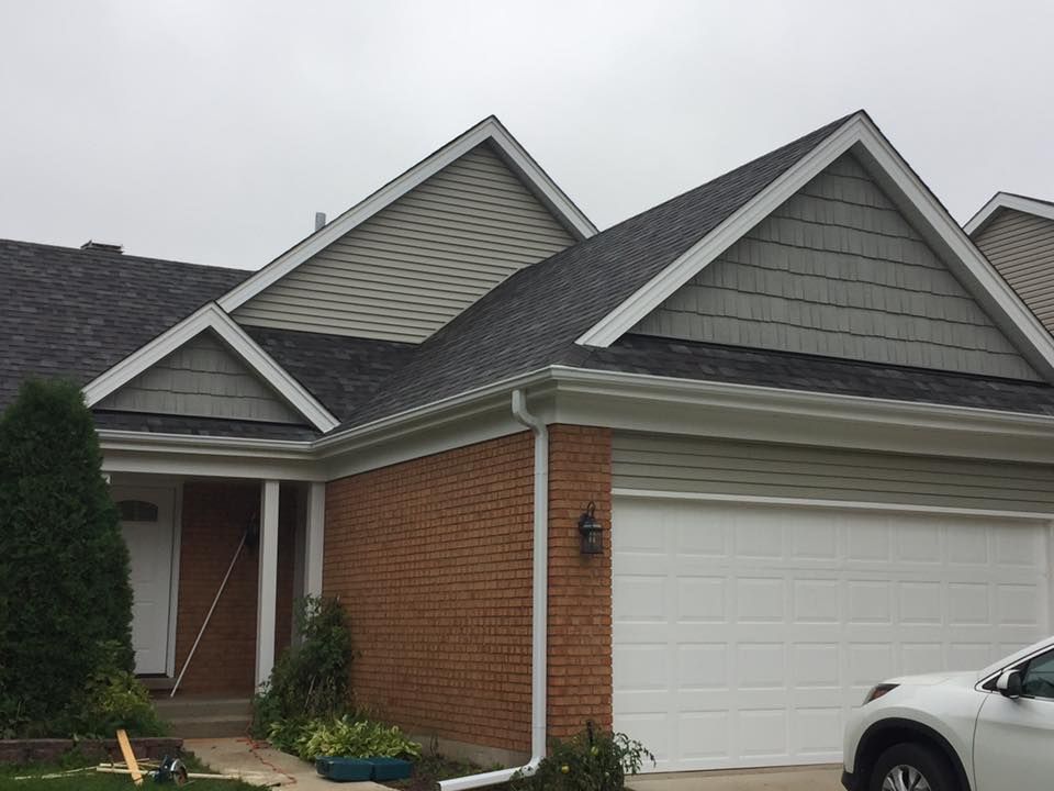 A suburban house exterior with a brick garage, white trim, grey shingled roof, and mixed siding gables on a cloudy day.