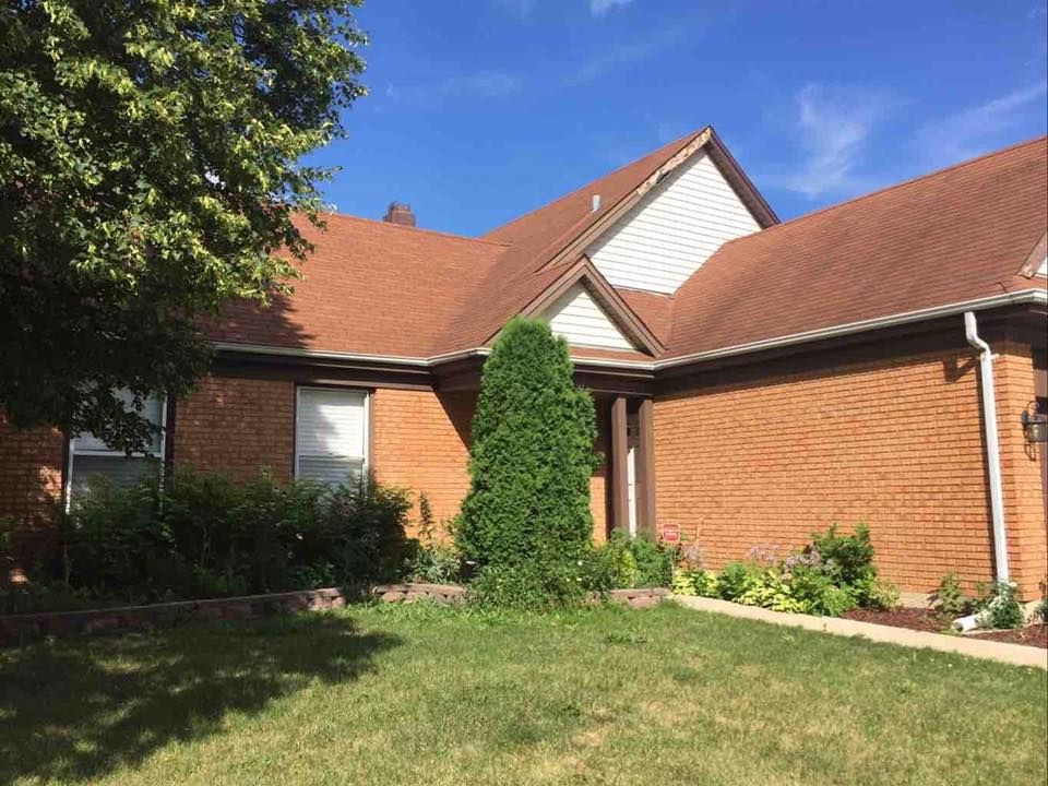 A one-story brick house with a brown shingled roof, white siding gables, and a green front yard under a blue sky.