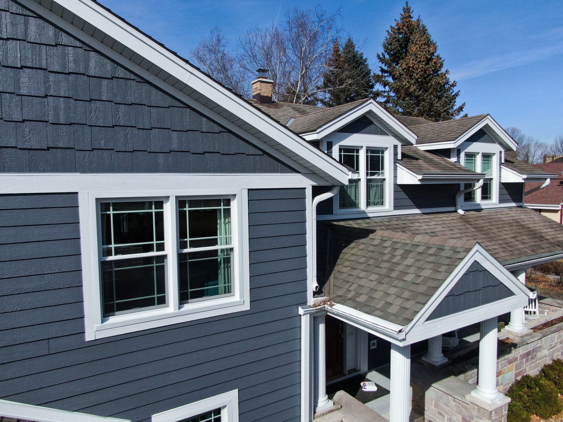 A two-story house with dark blue shingle siding, white trim, and a shingled roof under a clear blue sky.