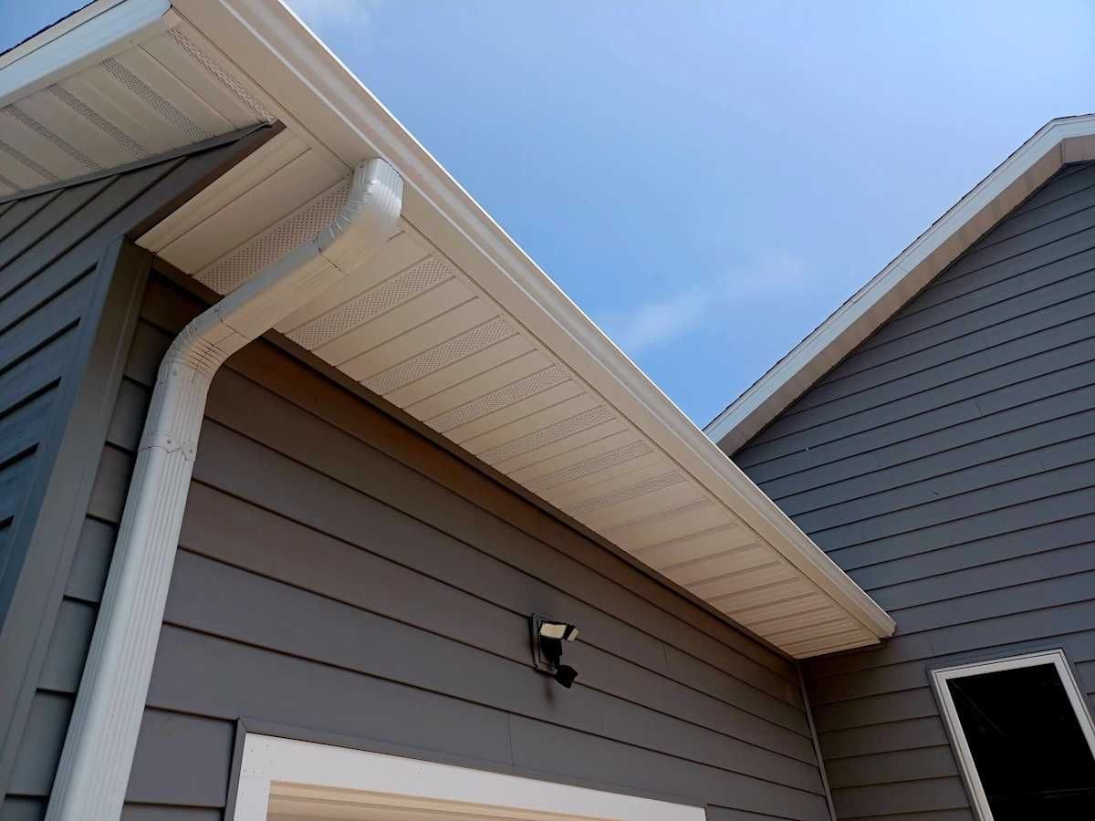 Low-angle shot of a house exterior showing a white gutter and soffit against gray siding under a clear blue sky.