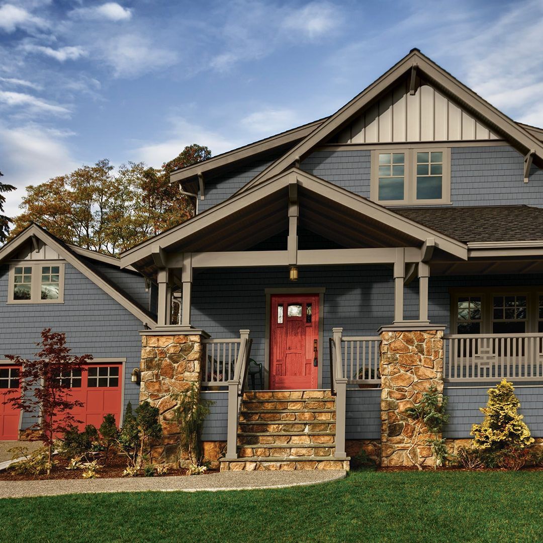 A two-story blue house with a stone foundation, a covered porch, and vibrant red doors set against a clear blue sky.