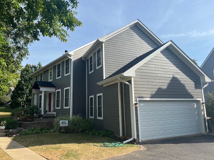 A two-story house with grey horizontal siding, white trim, a front porch, and an attached two-car garage.
