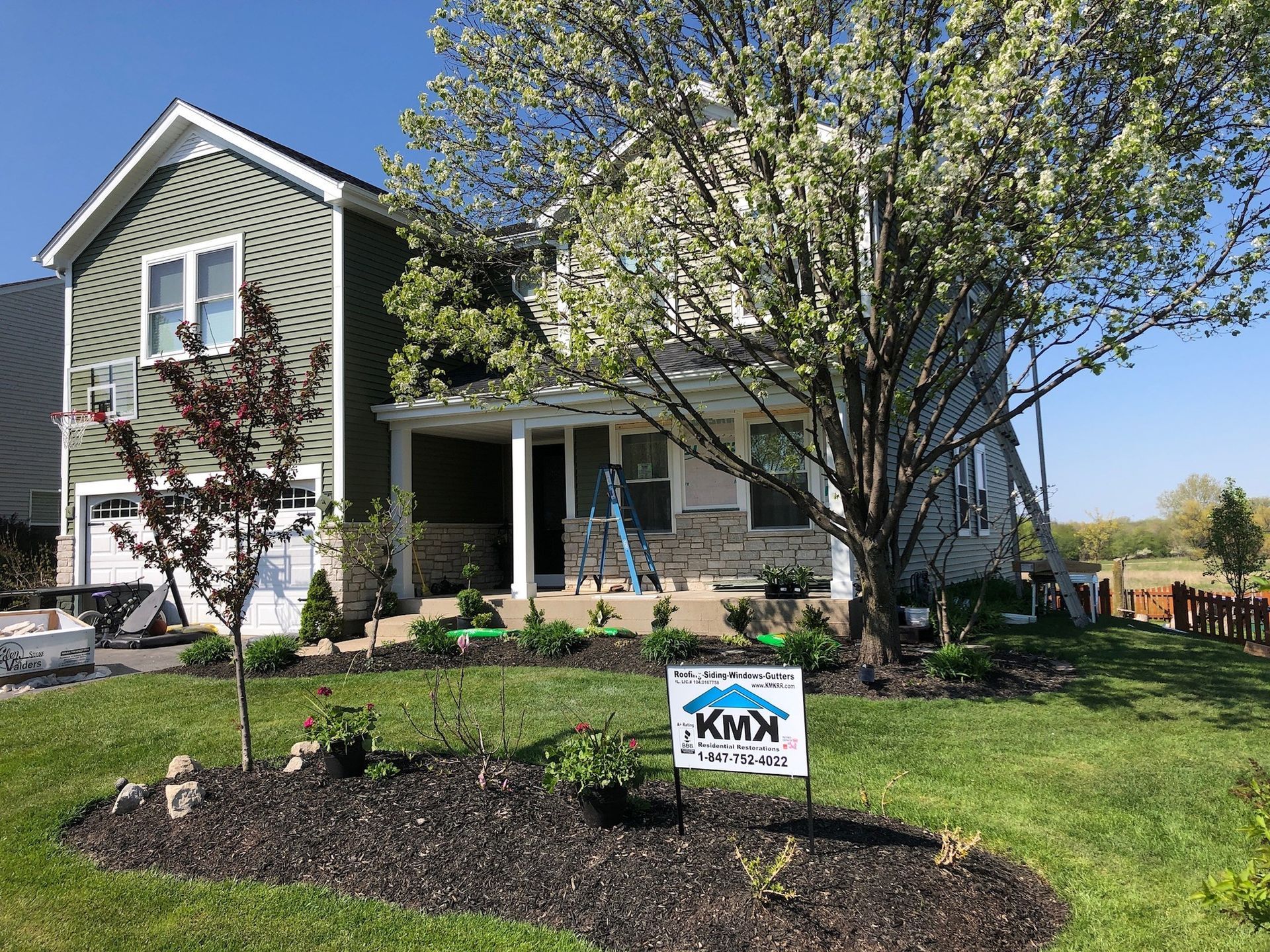 A two-story green-sided house with stone accents, a landscaped front garden, a blooming tree, and a KMK construction sign.