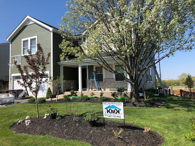 A green two-story suburban house with a covered front porch, a flowering tree, and a KMX Roofing sign in the front yard.