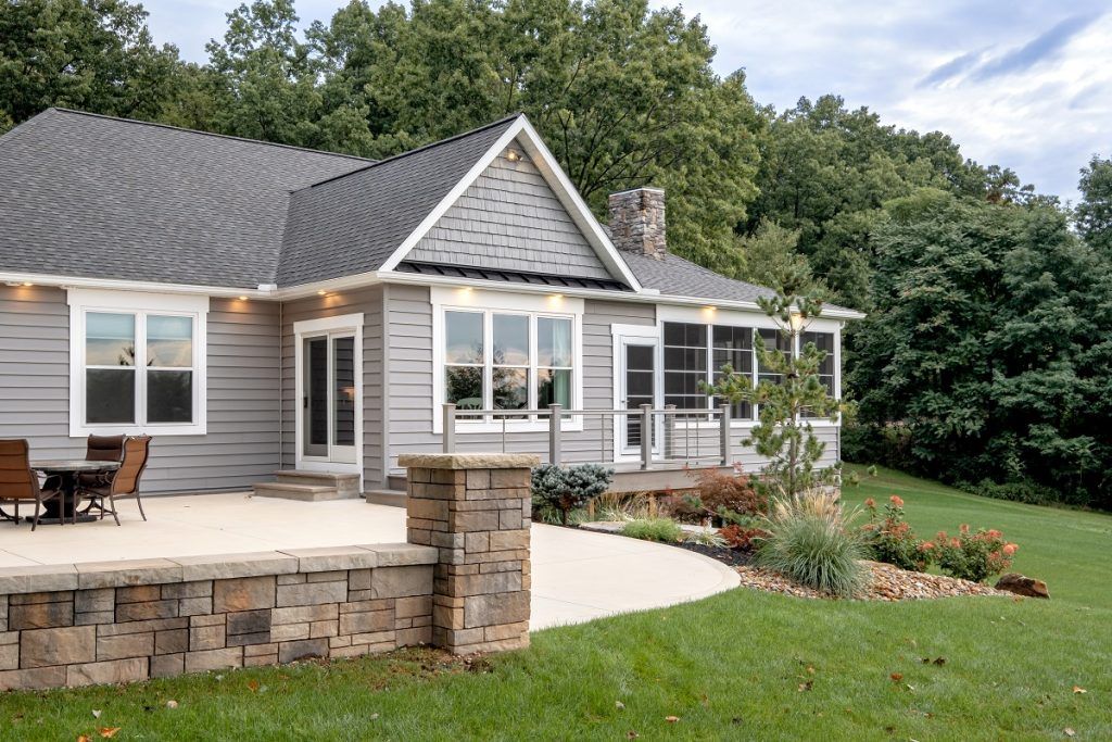 A gray house with a stone patio, low retaining wall, and landscaped lawn under a clear sky.
