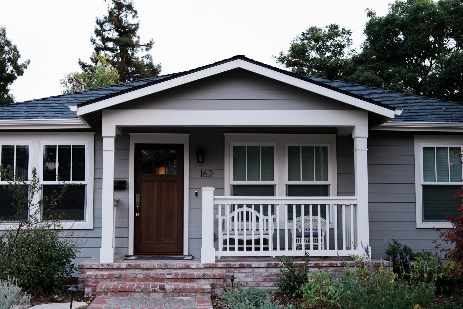 A gray, single-story house with a wooden front door, white porch railing, and red brick steps leading to the entrance.