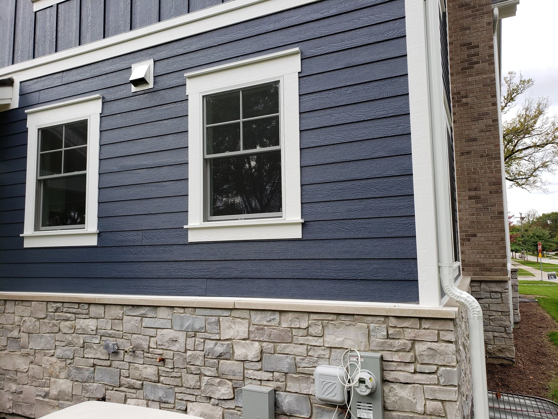 Dark blue shingle siding above a stone foundation on the exterior wall of a building with two white-trimmed windows.
