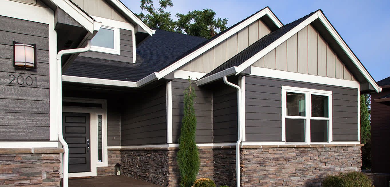 A modern suburban house exterior with gray horizontal siding, stone veneer base, white trim, and a dark front door.