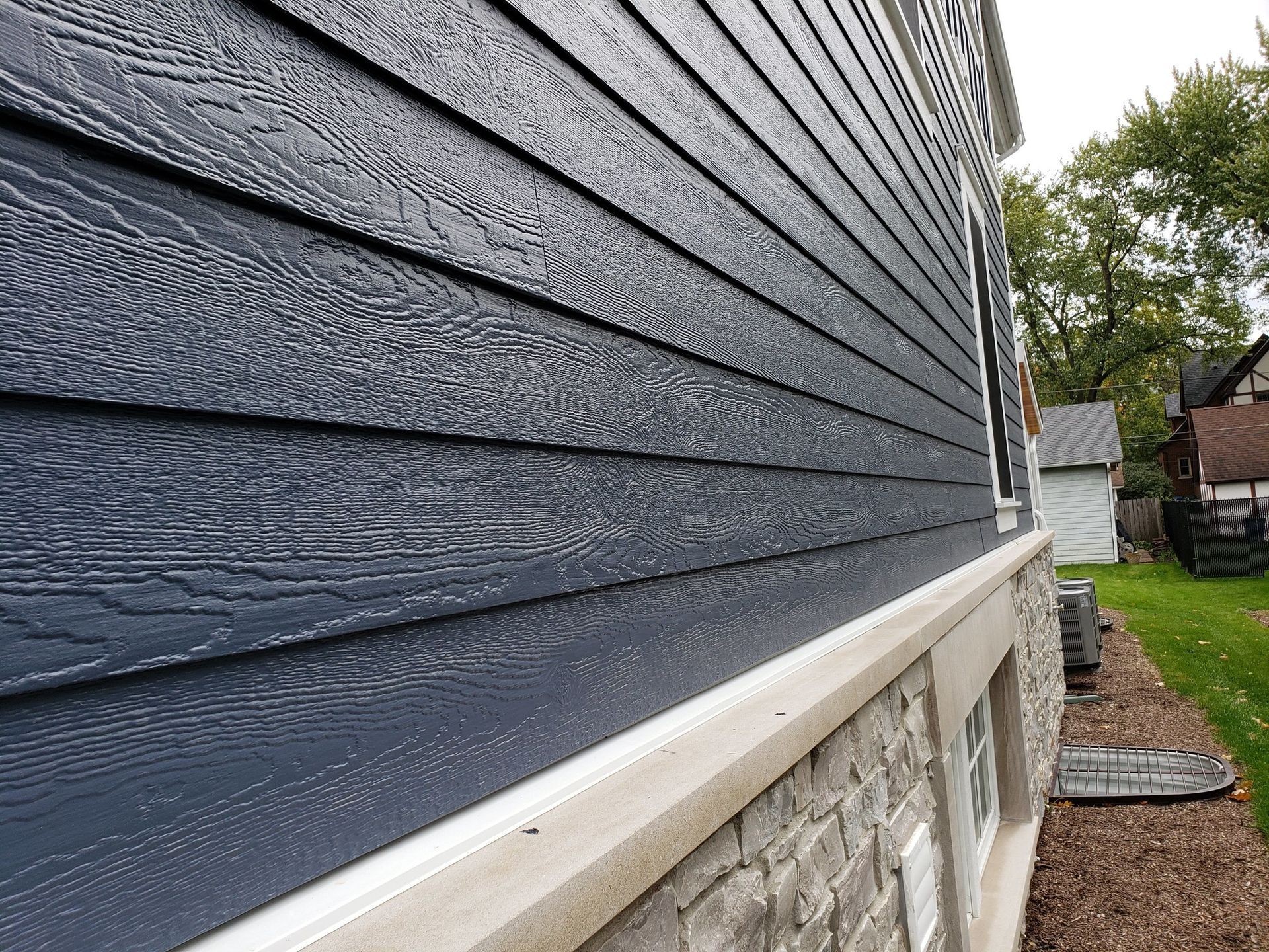 A side view of a house exterior with dark blue horizontal siding above a stone foundation, with trees in the background.