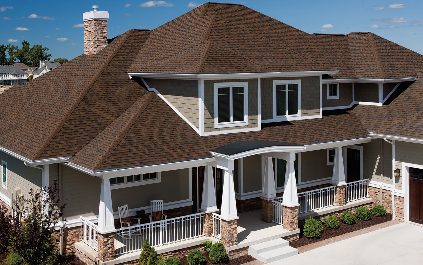 A two-story suburban home with tan siding, a brown shingled roof, a covered front porch, and stone accents.