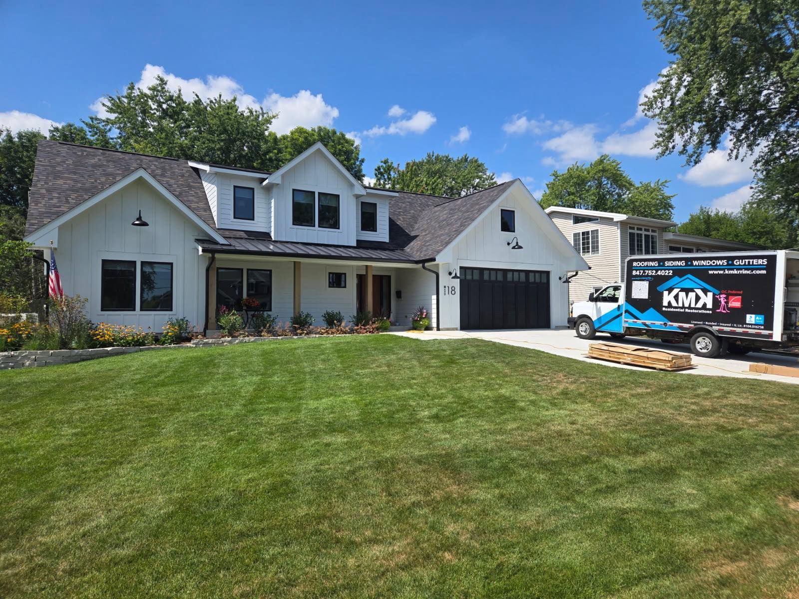 A newly renovated white farmhouse-style home with a dark roof and black garage, with a construction truck parked nearby.