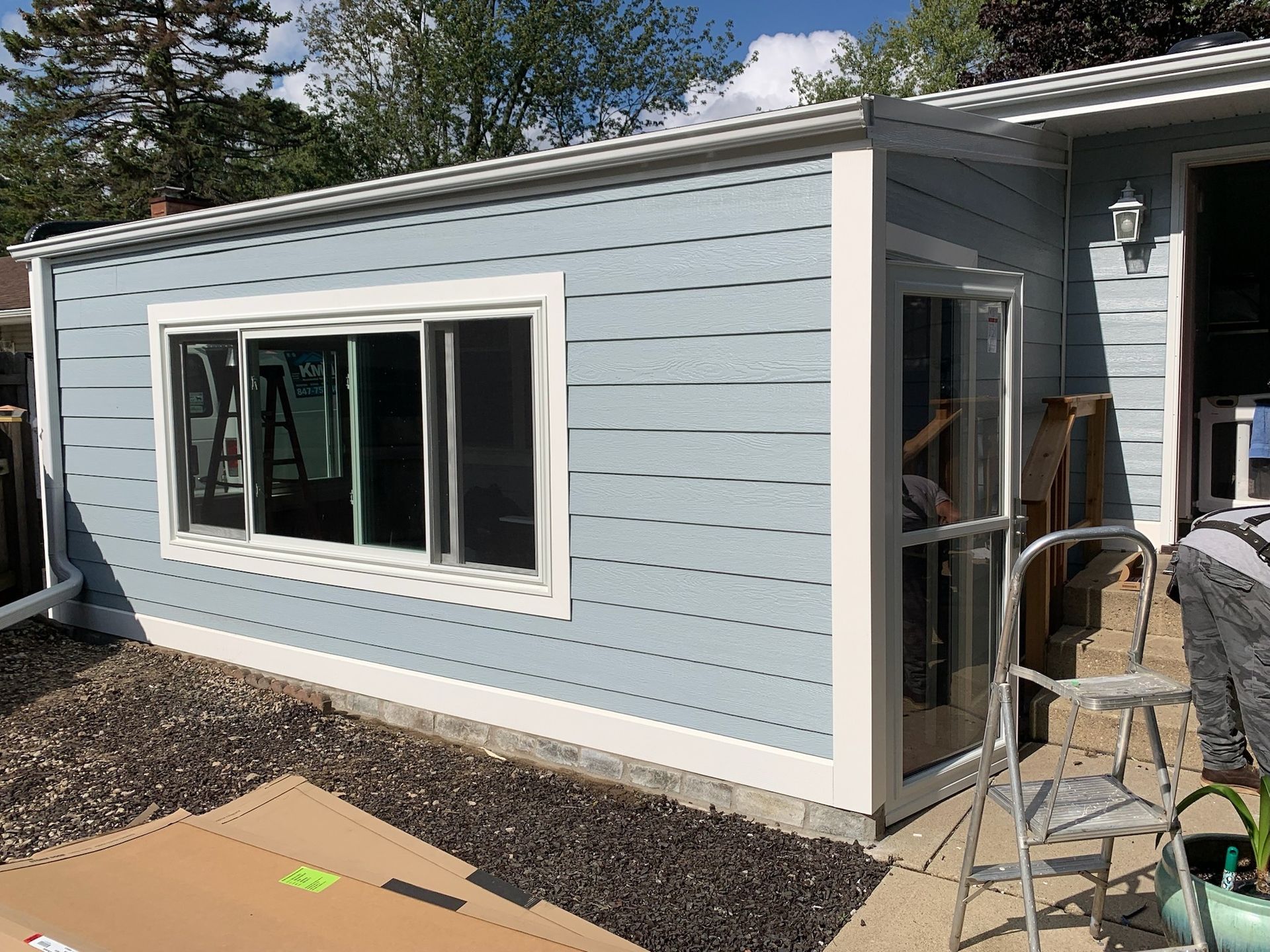 Light blue exterior wall of a home addition with a large sliding window, a side door, and a metal ladder on a patio.
