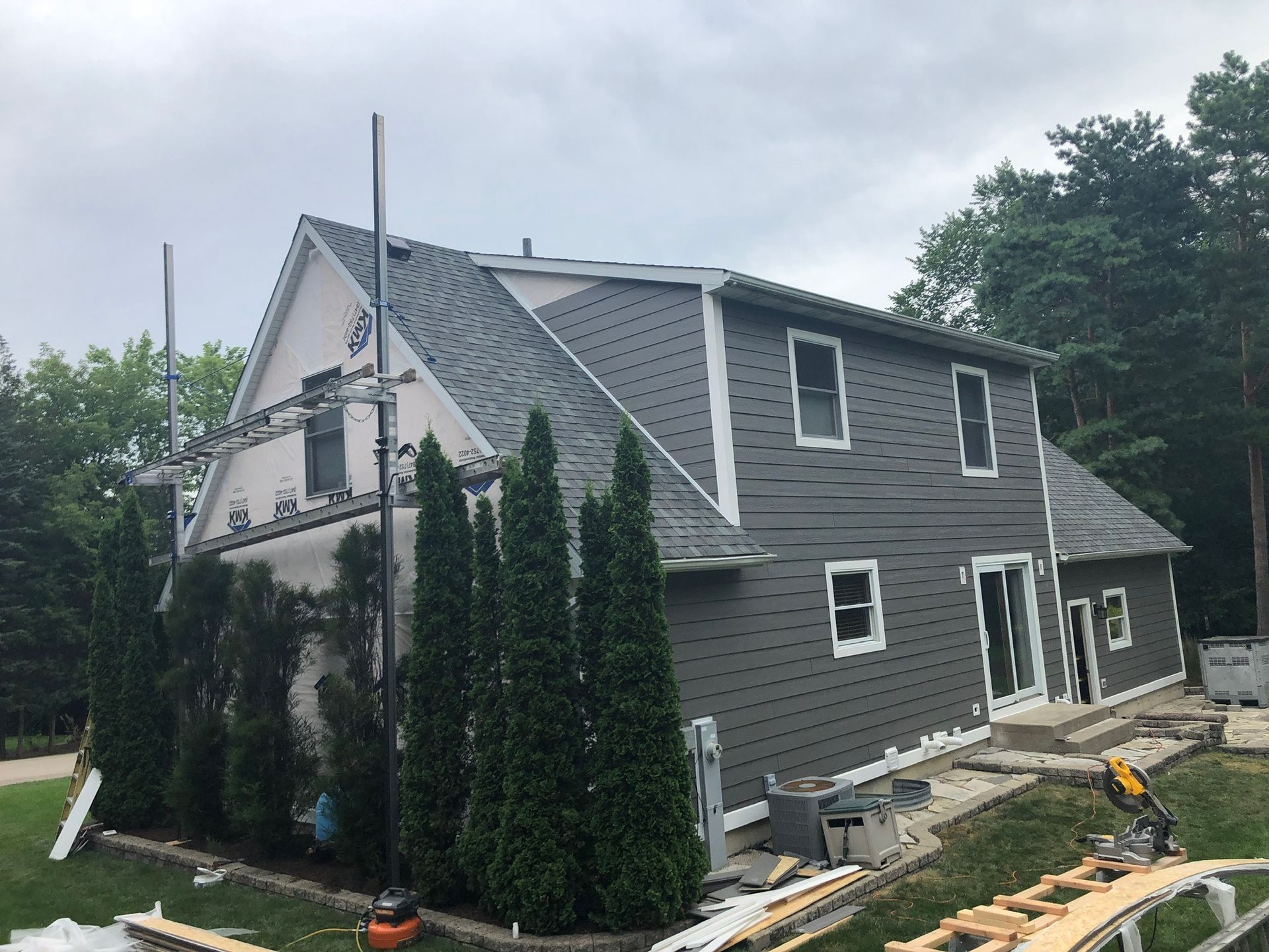 A gray-sided house under construction with scaffolding, surrounded by tall evergreen trees on a cloudy day.