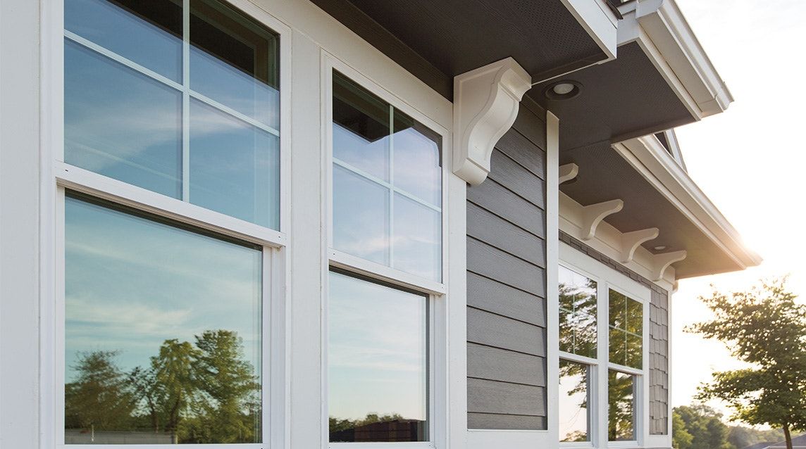 Low-angle view of gray horizontal siding on a house with white-framed double-hung windows and decorative roof brackets.
