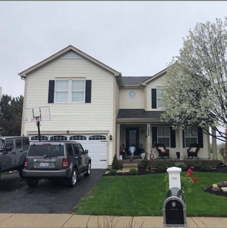 Two-story suburban house with cream siding, dark shutters, a front porch, a gray SUV in the driveway, and a blooming tree.