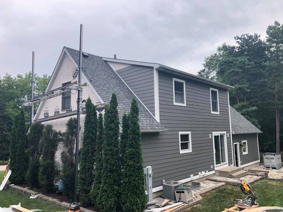 A house under construction with grey siding, a white-framed gable, scaffolding on the side, and pine trees in the yard.