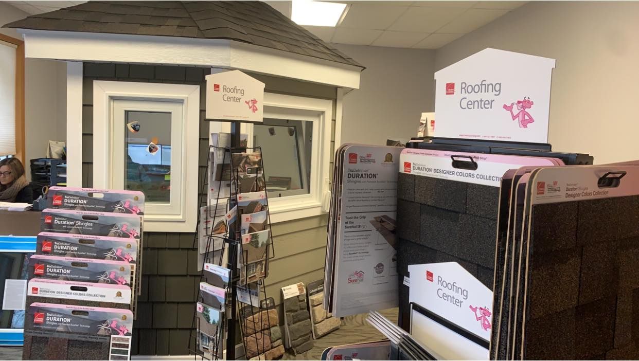 A showroom display featuring stacked roofing shingle samples, marketing signage, and a model house exterior.