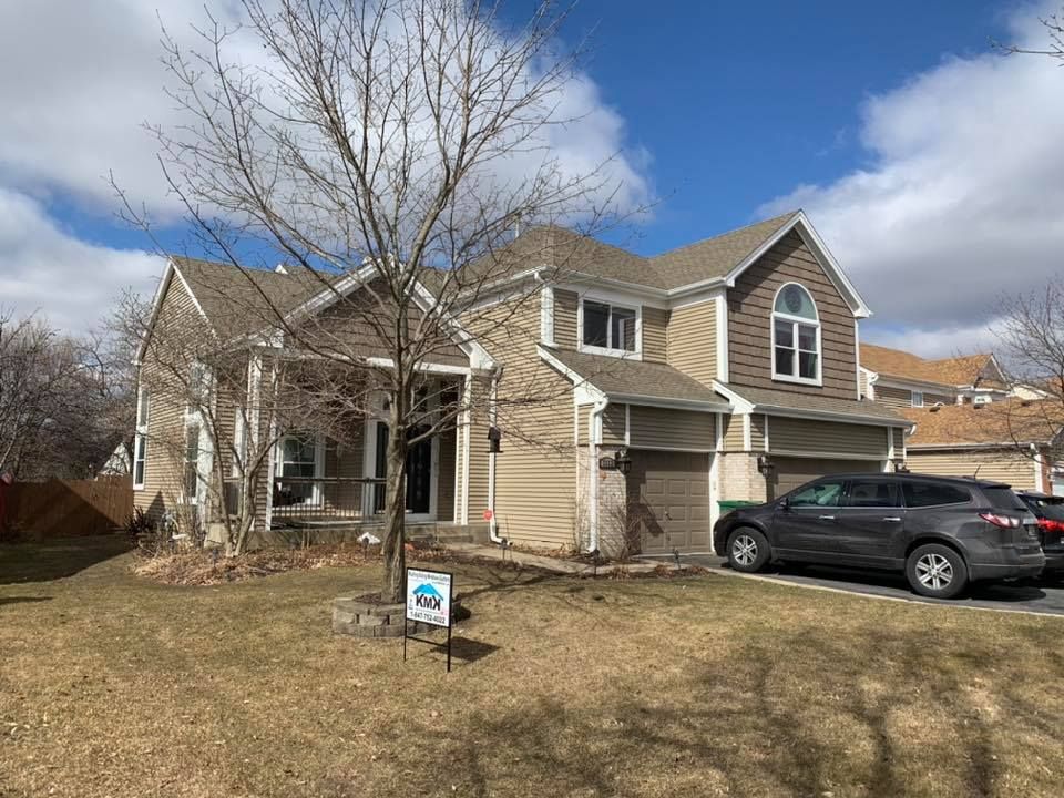 A tan, two-story house with a brown roof and a gray SUV parked in the driveway on a sunny day.